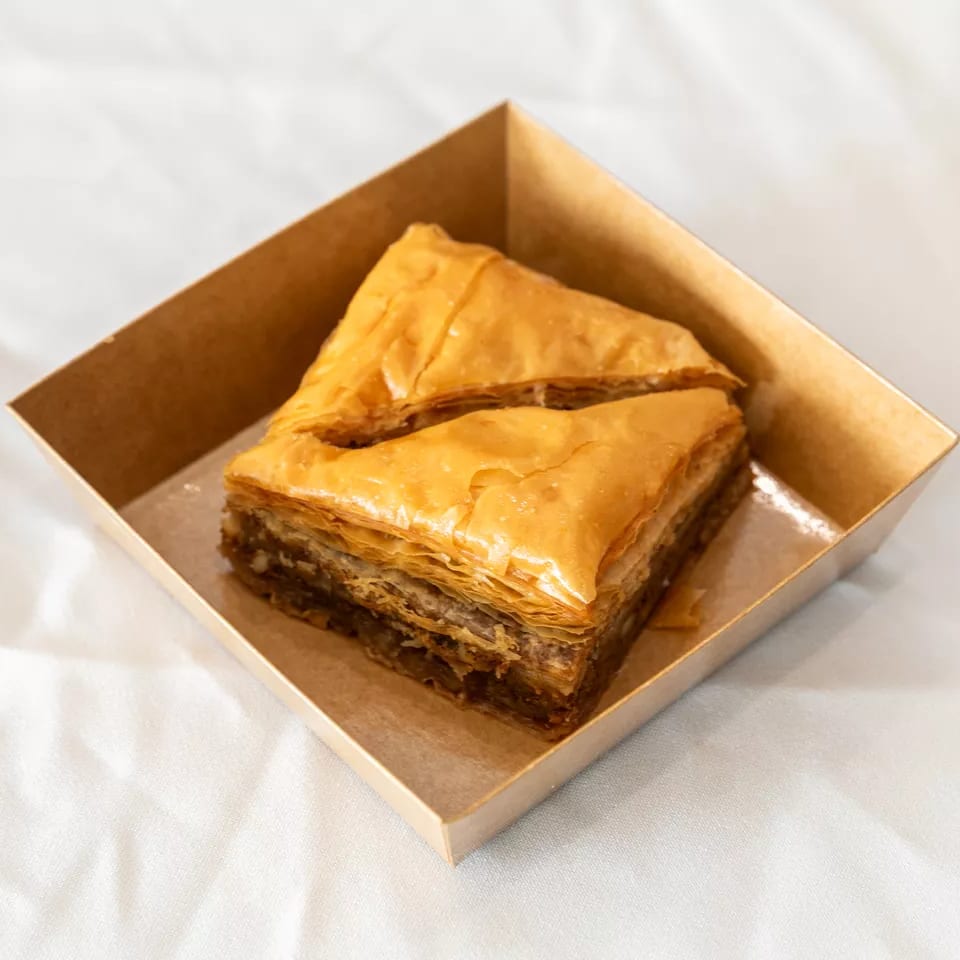 Two pieces of golden baklava in a brown cardboard container on a white tablecloth.