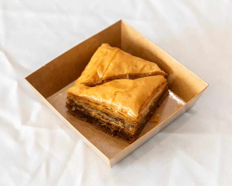 Two pieces of golden baklava in a brown cardboard container on a white tablecloth.
