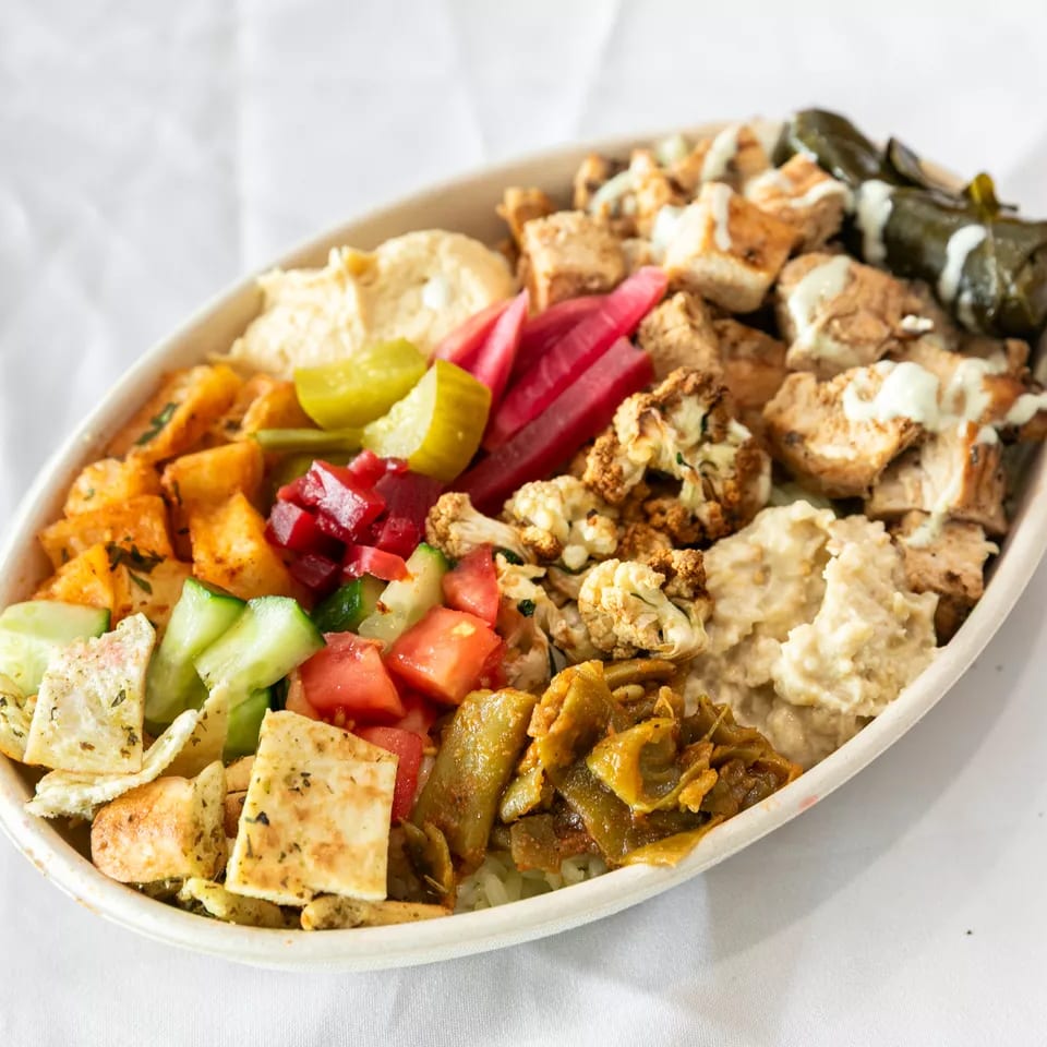 Colorful Mediterranean bowl with chicken, hummus, pickled vegetables, salad, and stuffed grape leaves.