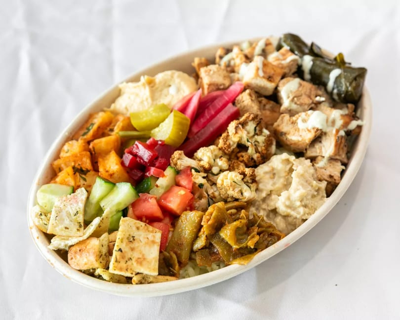 Colorful Mediterranean bowl with chicken, hummus, pickled vegetables, salad, and stuffed grape leaves.