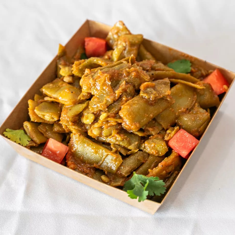 Stewed green beans and lentils in a rich sauce, garnished with tomatoes and cilantro in a takeaway box.