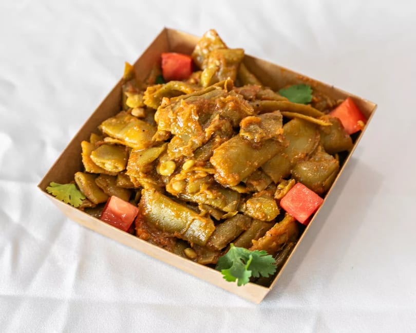 Stewed green beans and lentils in a rich sauce, garnished with tomatoes and cilantro in a takeaway box.
