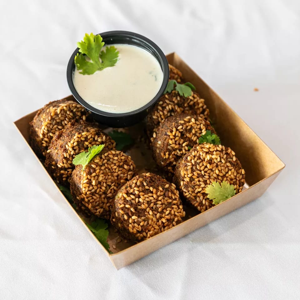 Sesame-crusted falafel balls in a cardboard tray with a side of white dipping sauce and cilantro.