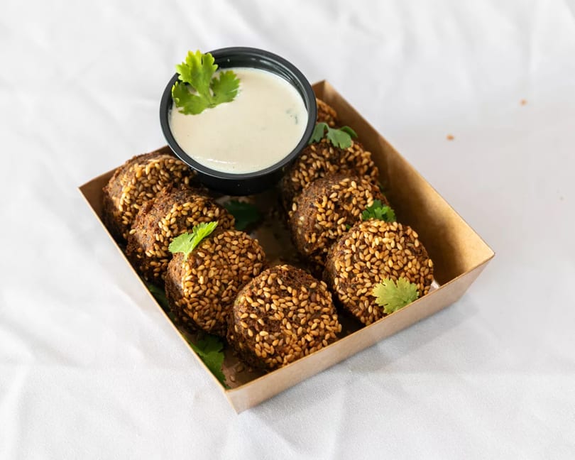 Sesame-crusted falafel balls in a cardboard tray with a side of white dipping sauce and cilantro.