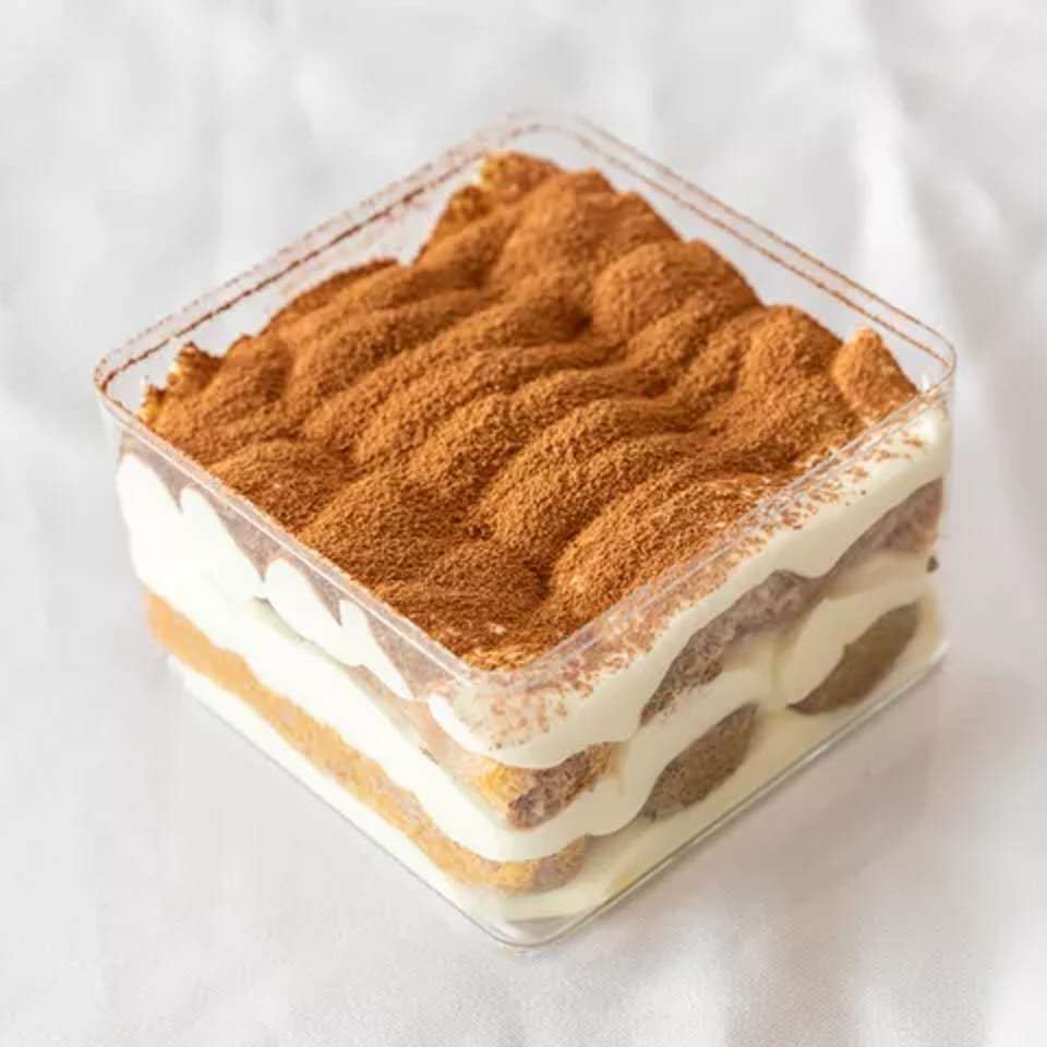 Layered tiramisu dessert in a clear square container, topped with cocoa powder.