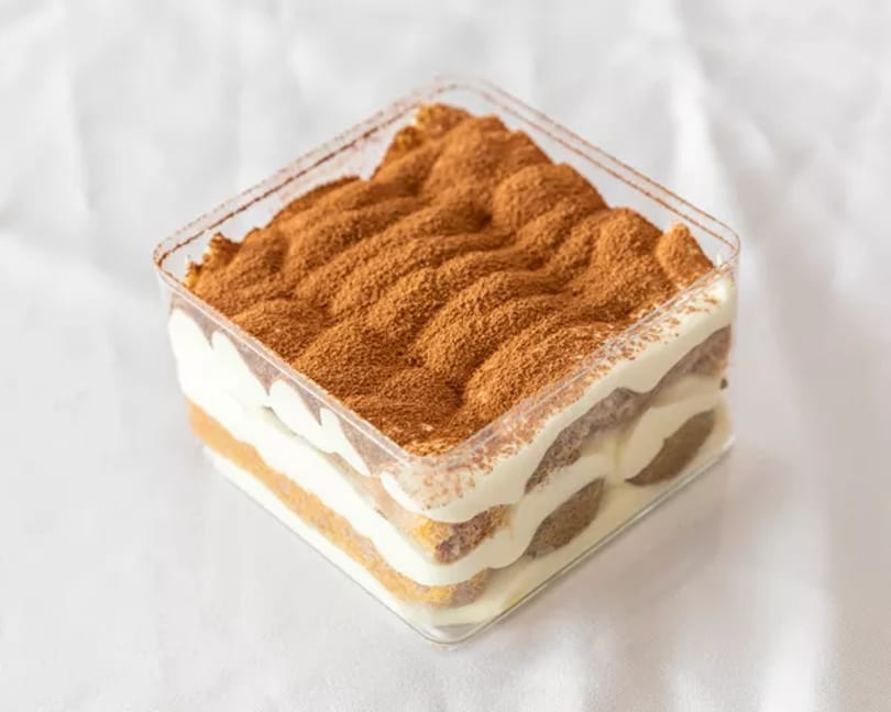 Layered tiramisu dessert in a clear square container, topped with cocoa powder.